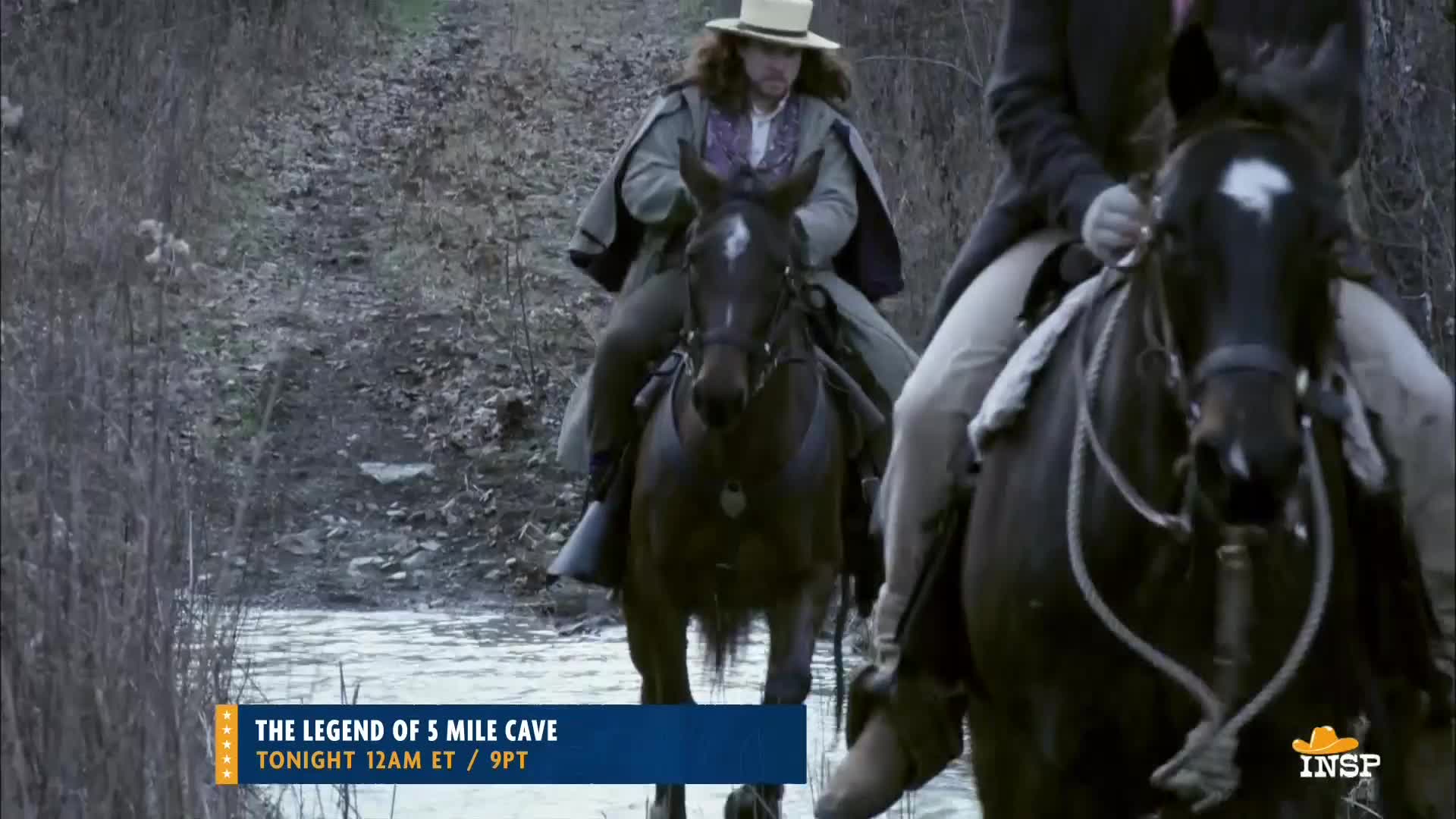 Two men on horseback ride through a shallow stream, their dark horses' coats reflecting the overcast sky. The lead rider, wearing a hat and cape, guides his mount forward.
