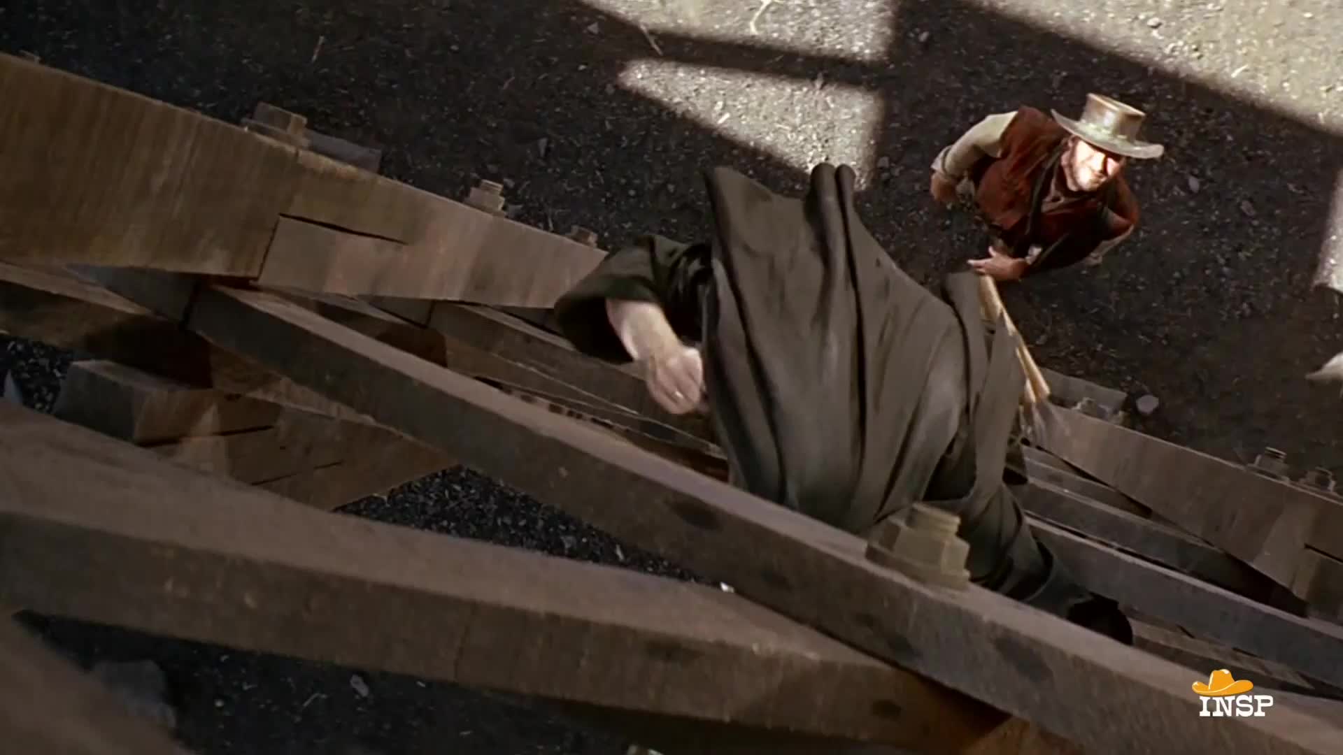 A man in a long coat struggles to climb across the wooden beams of a bridge. Another man, hat on his head, leans over him, seemingly about to strike with a wooden tool.
