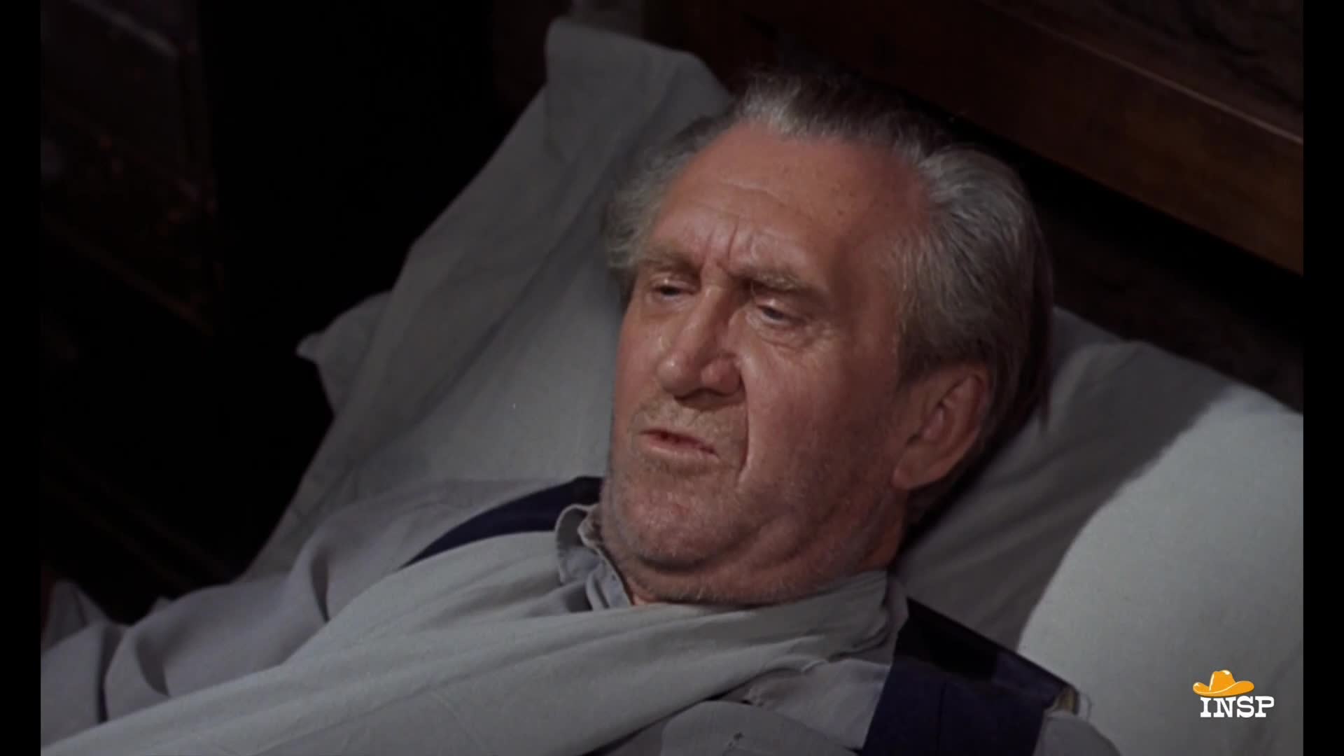 A man with graying hair lies in bed, his face creased with worry. He's wearing a light-colored shirt and suspenders, visible against the white sheets.
