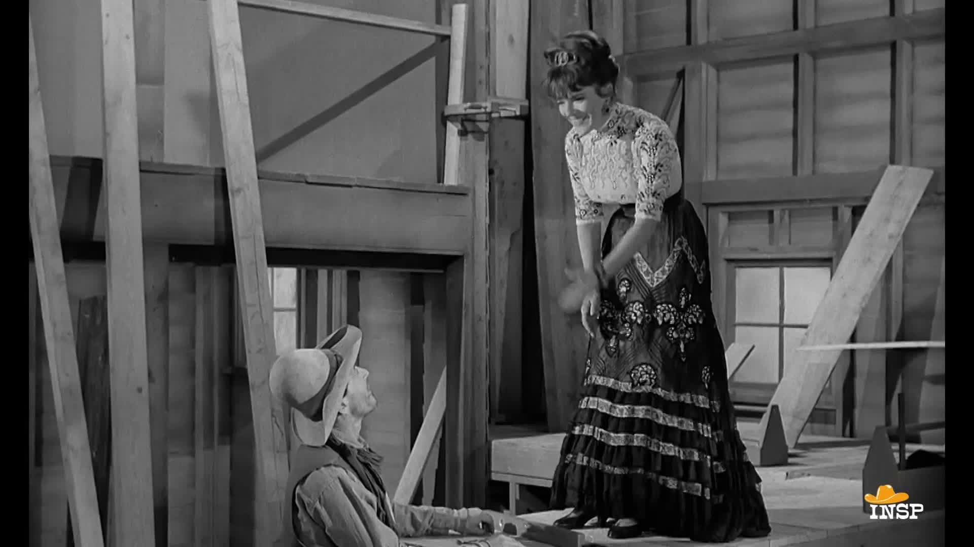 A woman in a long skirt laughs, looking down at a man in a cowboy hat. He's sitting on the floor, gazing up at her with a smile.

