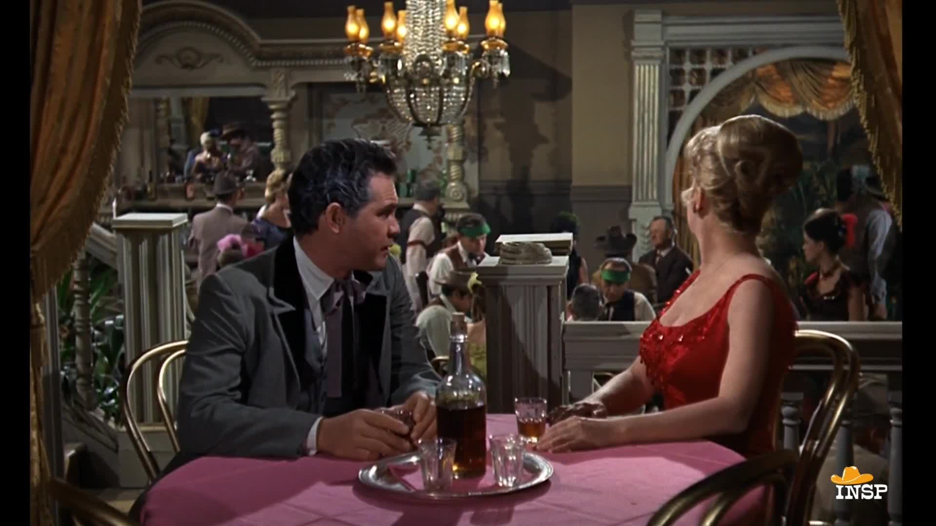 A man in a dark suit leans in, speaking to a woman in a red dress, as they sit at a table in a saloon. Behind them, other patrons and a bar are visible, lit by a large chandelier.
