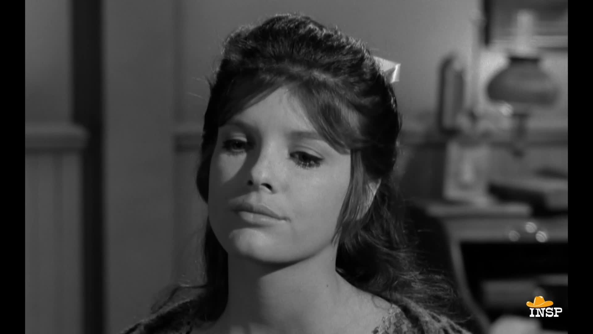 A woman with dark hair and a white bow sits, her gaze lowered. The scene, broadcast by INSP, is a close-up, likely from a Western.
