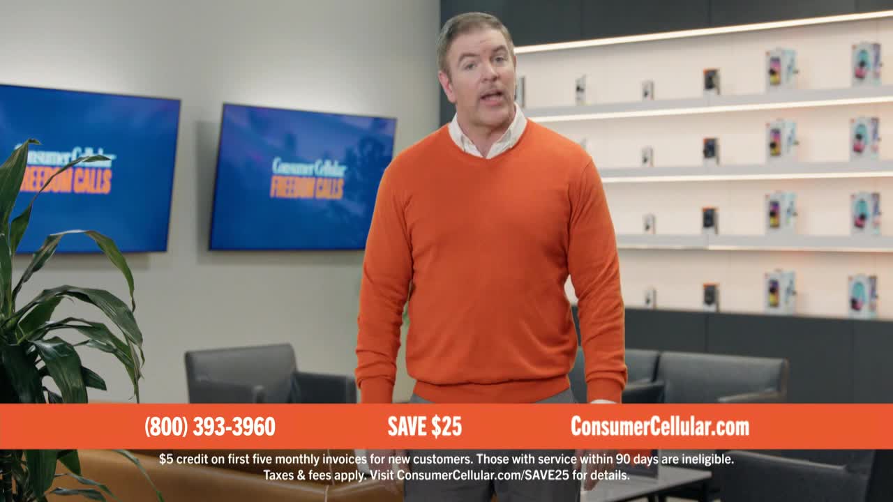 A man in an orange sweater stands in a modern office, talking to the camera. Behind him, two screens display the Consumer Cellular logo, and a shelf showcases phones.

