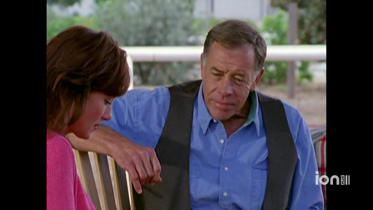 A man in a blue shirt and a dark vest leans toward a woman, his expression somber. She's in a pink sweater, looking down, and resting her hand on the bench.
