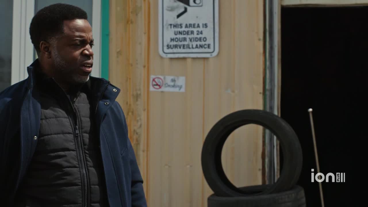 A man in a dark coat looks off to the side, his expression concerned. Behind him, a sign warns of video surveillance, and a stack of tires sits nearby.
