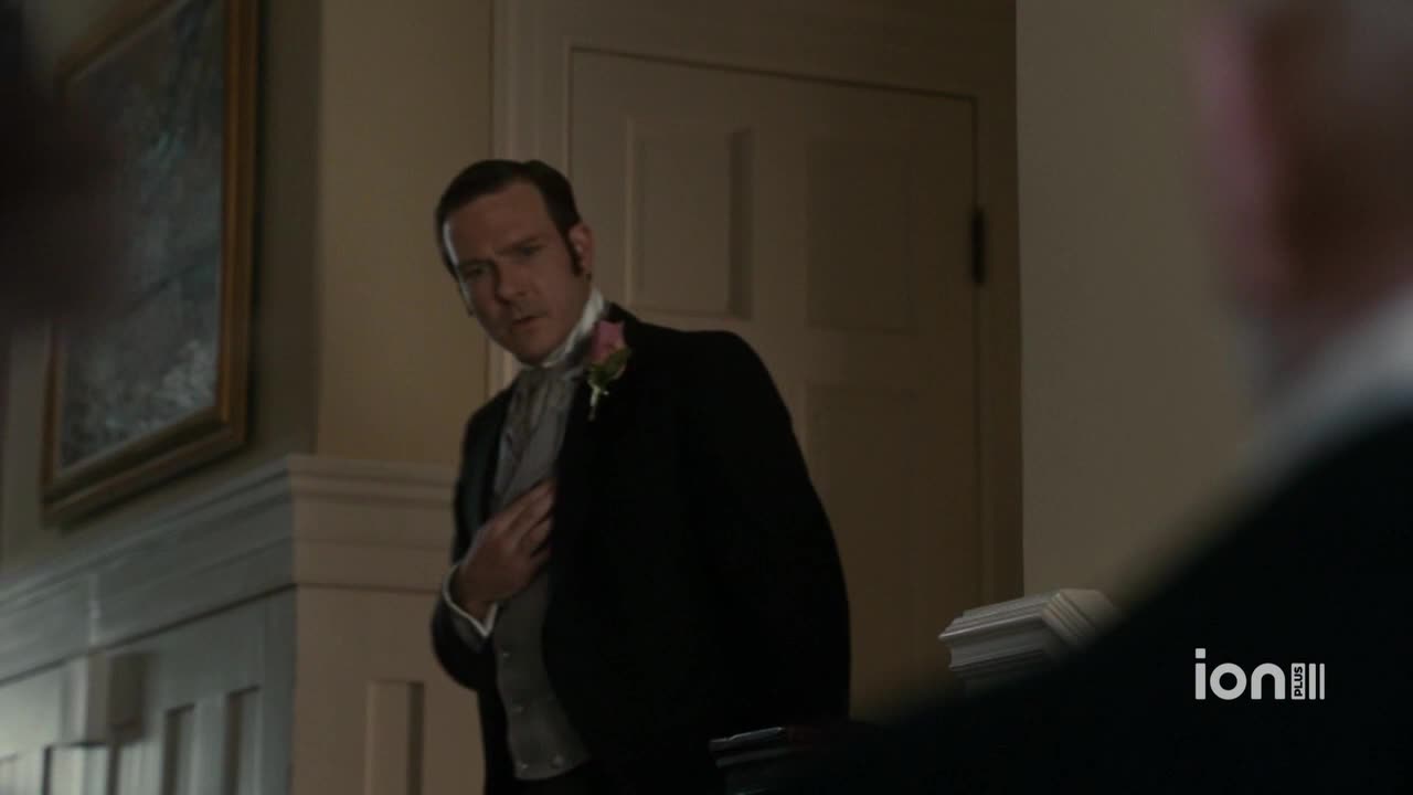 A man in a formal suit adjusts his waistcoat, a pink flower pinned to his lapel. He looks toward someone out of frame, his expression a mixture of concern and curiosity.
