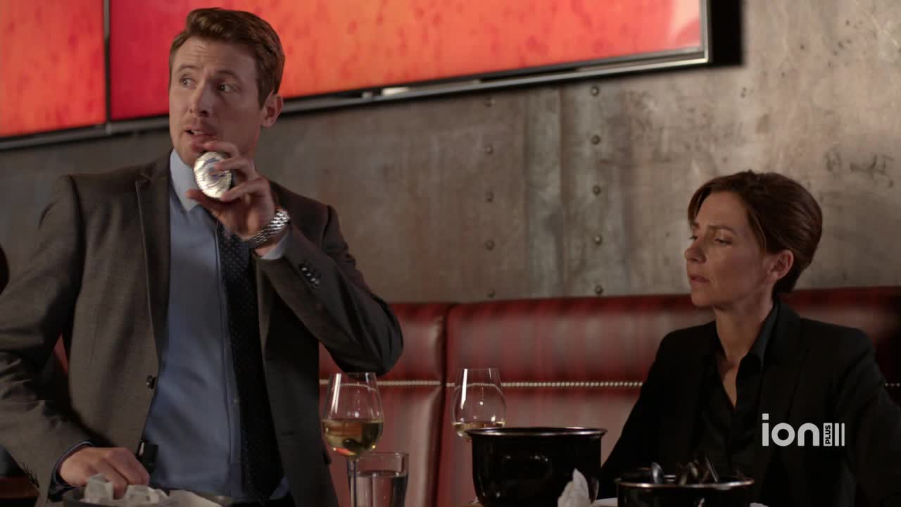 A man in a suit holds a small, round object up to his mouth. Across the table, a woman in a blazer sits quietly, looking down.
