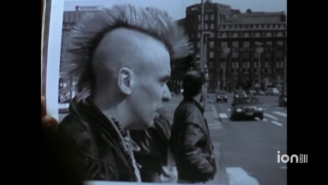 A picture of a man with a mohawk is being held up, obscuring the foreground. Across the street, cars move slowly in front of a tall building.
