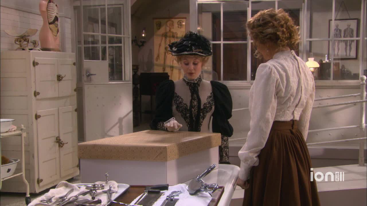 A woman in a black hat gestures toward a surgical table, while another in a white blouse listens intently. Surgical instruments gleam on a tray in the foreground, and a pink anatomical model stands nearby.
