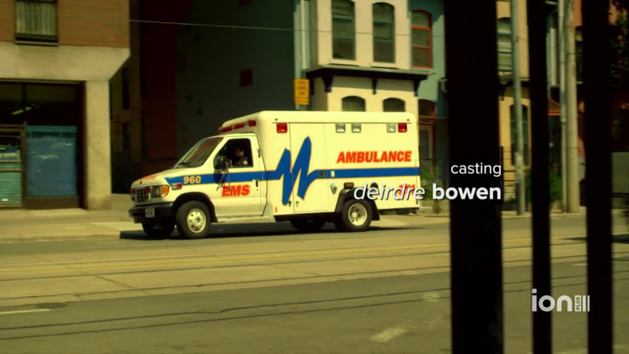 An ambulance, its lights off, rolls down the street. The vehicle is white with blue and red accents, and the word "AMBULANCE" is clearly visible.
