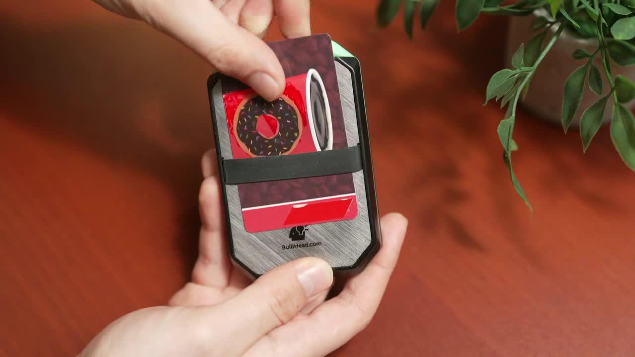 A hand is sliding a card out of a metal wallet, revealing a picture of a donut. The wallet is held in another hand, resting on a wooden surface.
