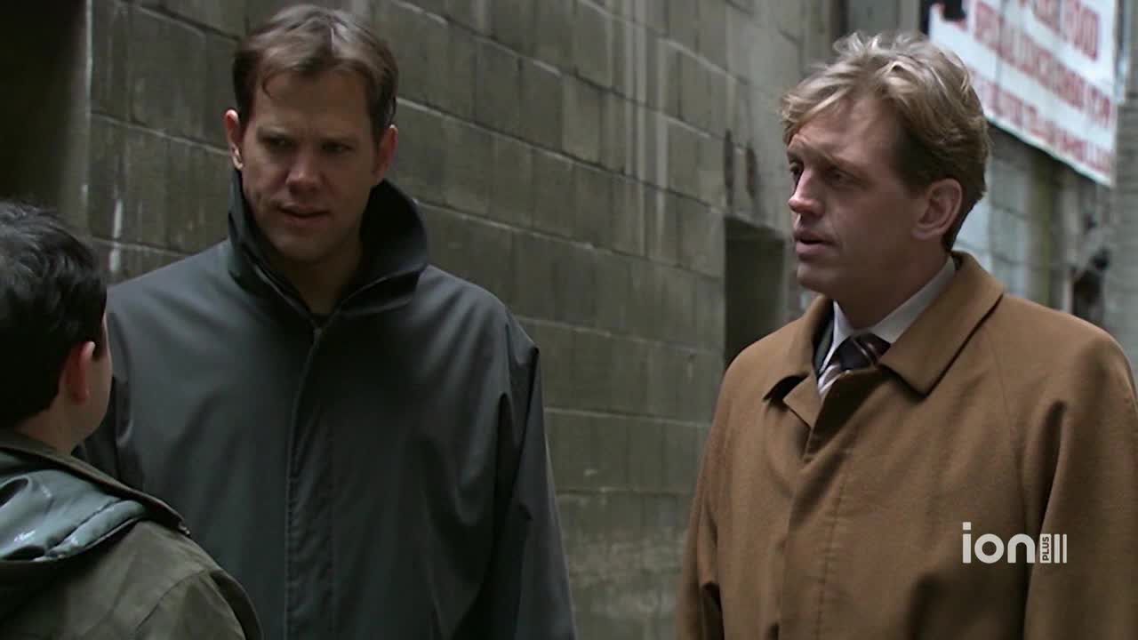 Two men in coats stand in a narrow alley, listening intently to a younger boy. The man on the right, wearing a tan coat, seems to be responding to the boy's words.
