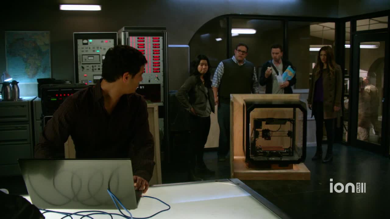 A man at a laptop turns to look at a group gathered around a large, enclosed machine. The ION Plus logo is visible, and the scene appears to be from a show set in the United States.
