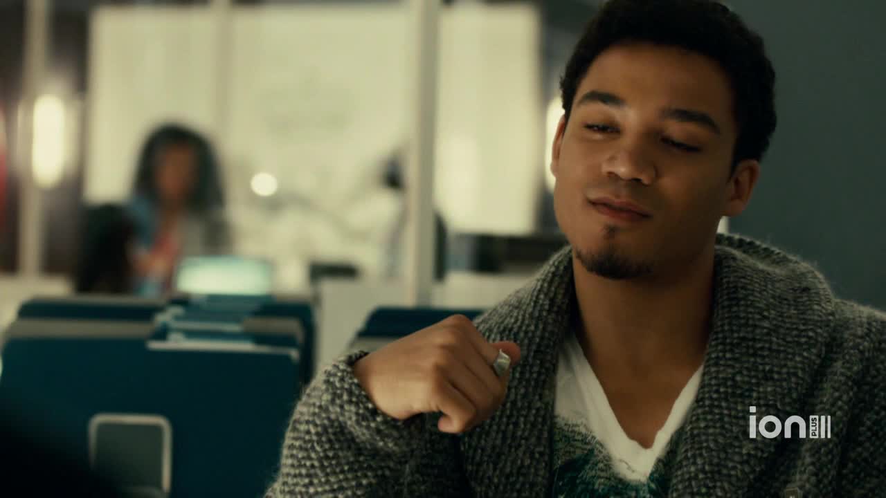 A man in a textured gray sweater leans forward, a slight smile playing on his lips. Behind him, a blurry figure sits at a desk in what appears to be an office setting, as seen on ION Plus.
