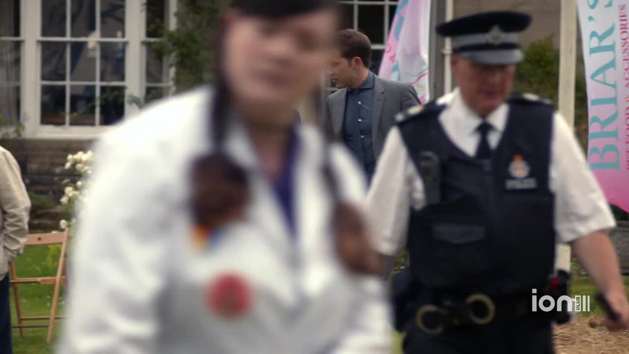 A police officer in a dark vest and hat walks toward the right, his gaze forward. A man in a suit jacket stands nearby, while a doctor in a white coat is in the foreground.
