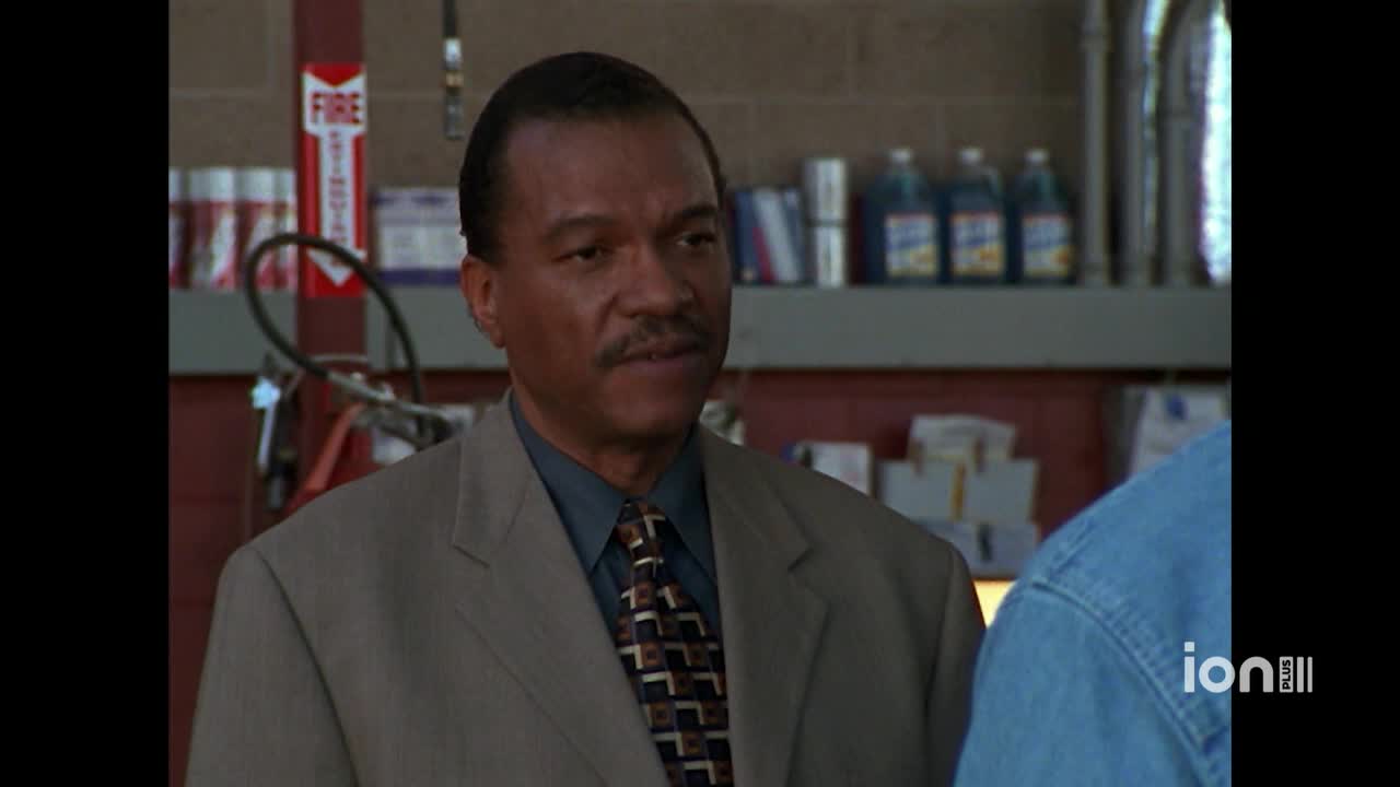 A man in a suit and tie is talking to someone out of frame, his expression thoughtful. Behind him, a fire extinguisher and some cleaning supplies sit on a shelf, as seen on ION Plus.
