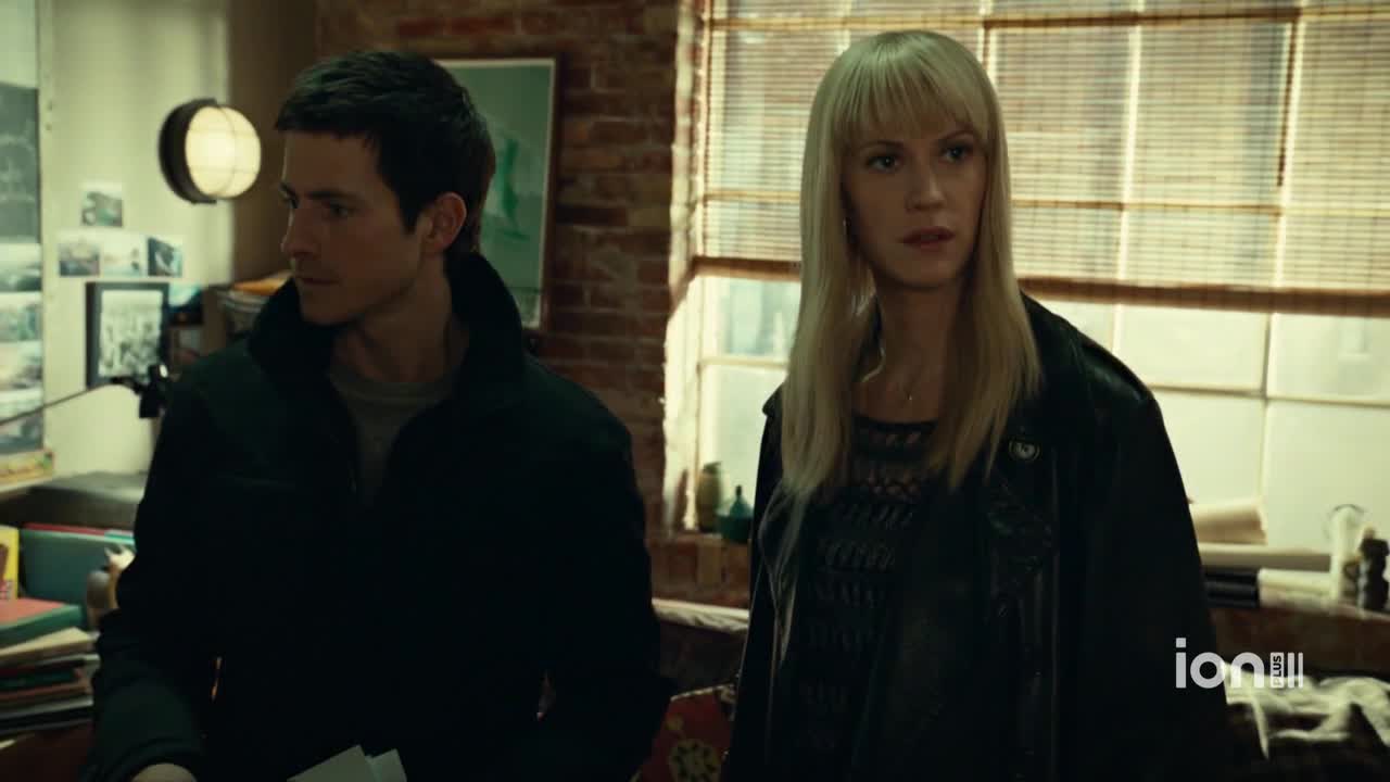 A man in a dark jacket glances to his left while holding a stack of papers. Beside him, a woman with blonde bangs and a black leather jacket looks forward, her expression serious.
