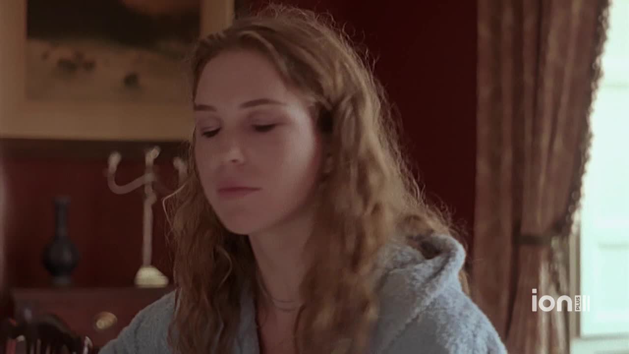 A woman with long, wavy hair is looking down, her expression thoughtful. She's wearing a light blue robe and seems to be in a room with dark red walls, as seen on ION Plus.
