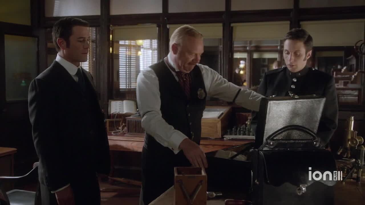 A man in a vest and badge opens a black bag, revealing its contents to two other men. The man on the right leans in, peering intently at whatever is inside.
