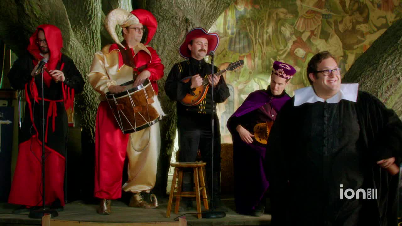 A costumed band, possibly for an ION Plus show, is set up on a stage. The musicians, dressed in medieval garb, are playing instruments and singing into microphones.

