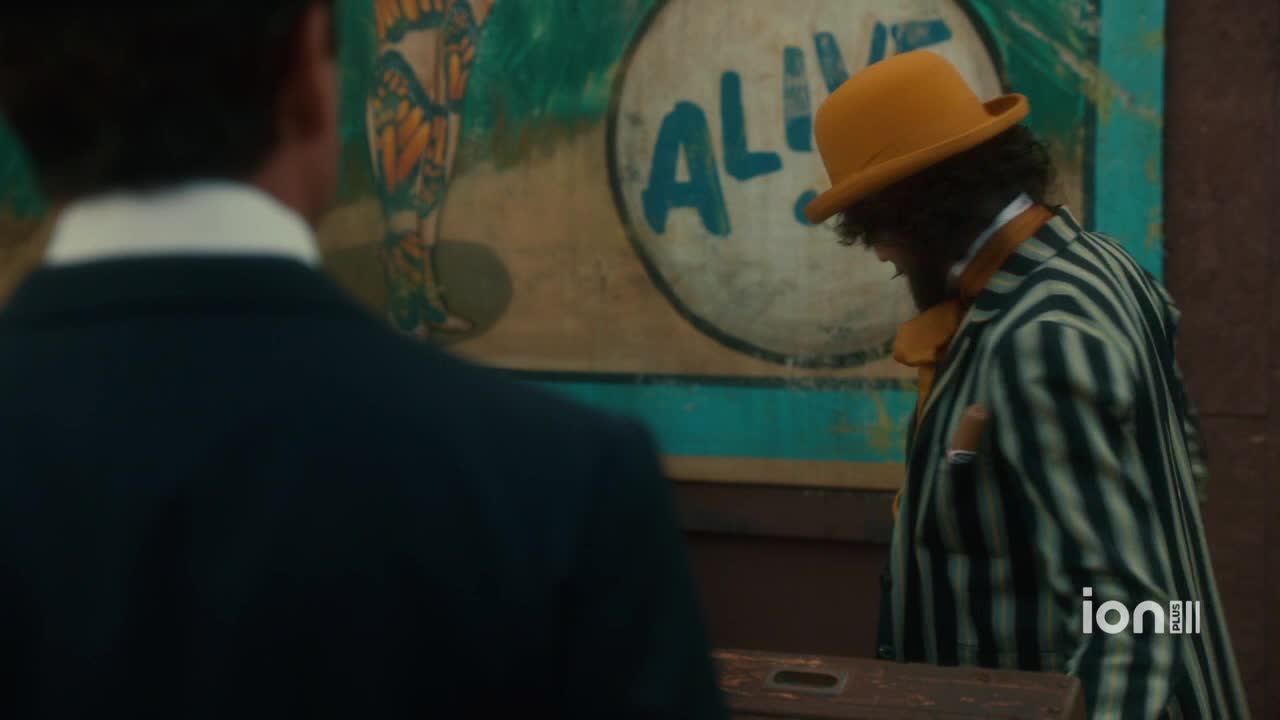 A man in a striped suit and a yellow hat stands near a painted sign that says "ALIVE." He seems to be looking down, as if contemplating something.
