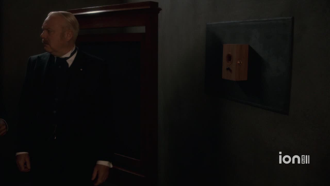 A man in a dark suit glances to his left, his face lit by an unseen light. A small wooden device is mounted on the wall to his right.
