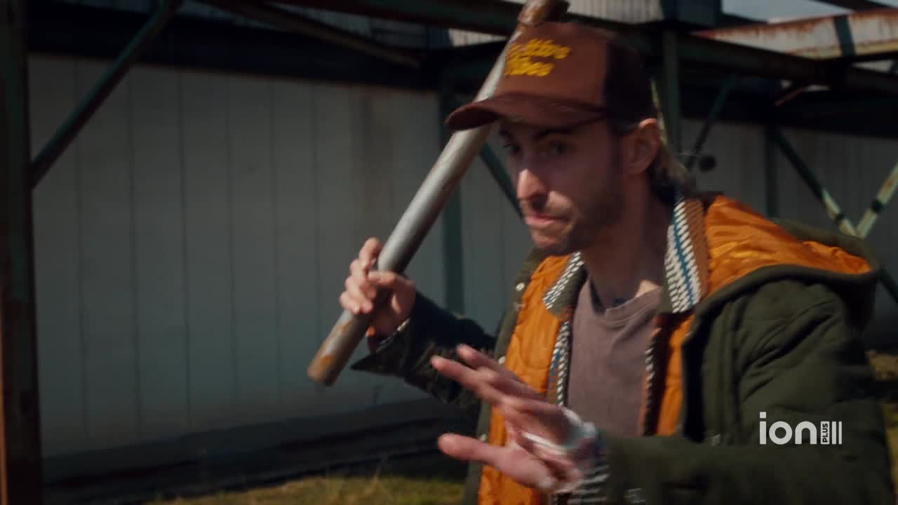 The man in the brown cap holds a metal pipe, gesturing with his other hand. He's wearing a green jacket with an orange lining, standing outside near a large building.
