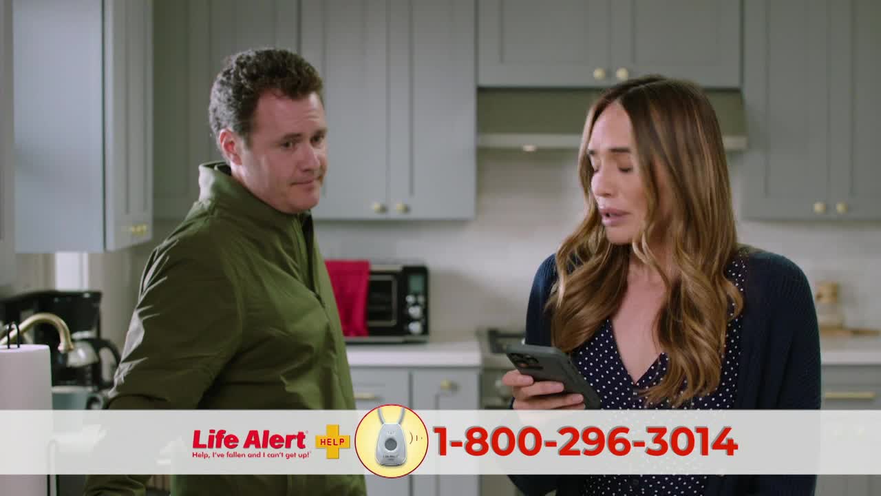 A woman in a blue sweater looks at her phone in a kitchen, while a man in a green jacket glances over.  A Life Alert commercial is playing on ION Plus.
