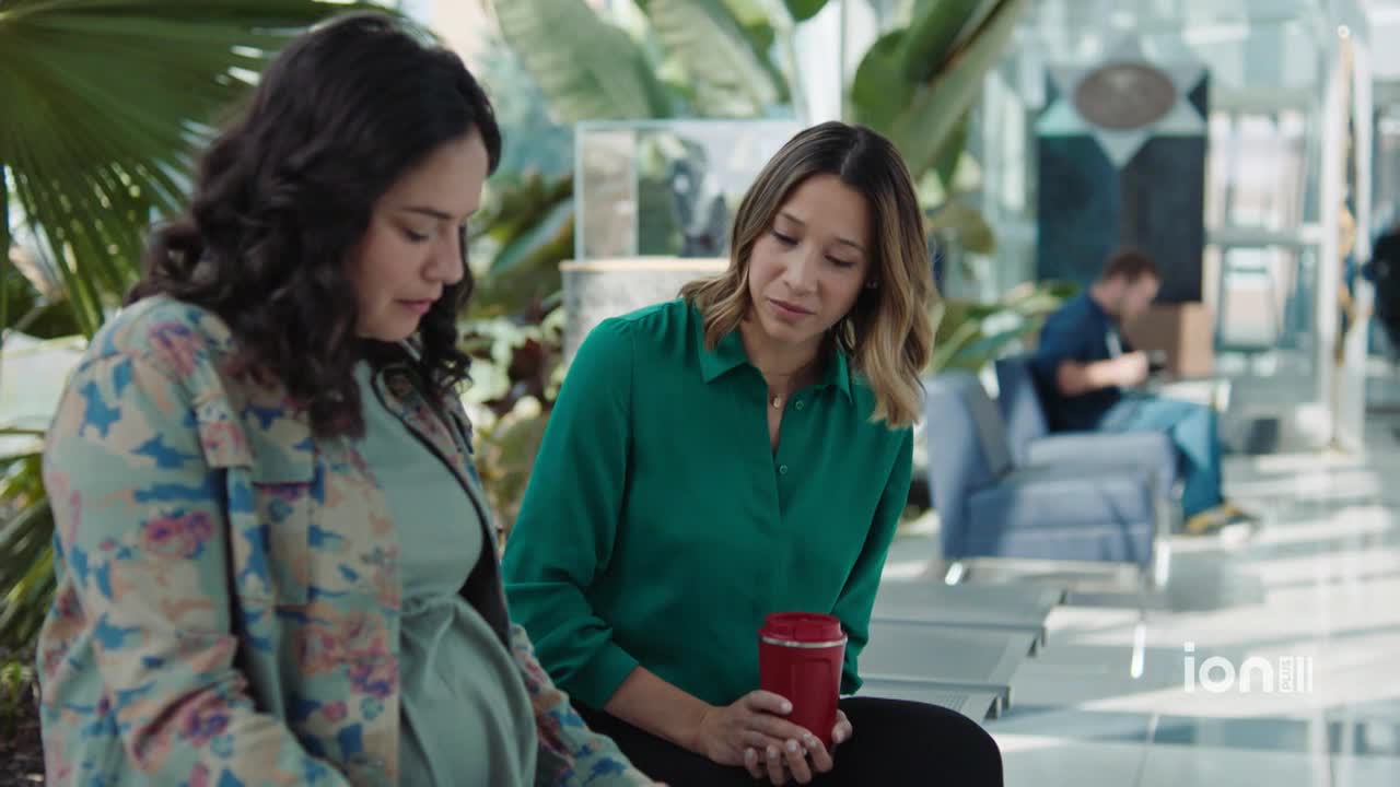 A pregnant woman in a floral jacket sits next to another woman holding a red travel mug. They're both looking down, seemingly focused on something out of view.
