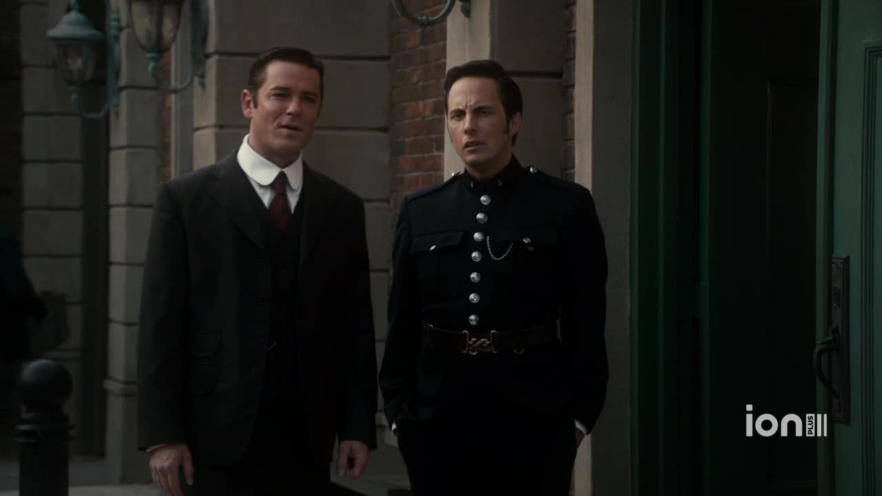 Two men stand outside, one in a dark suit and the other in a police uniform. The man in the uniform seems to be listening intently, while the other speaks.
