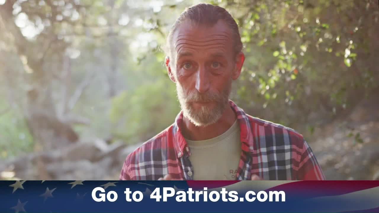 A man in a plaid shirt, with a trimmed beard, looks directly at the camera. Beneath him, a banner with the American flag and a website address appears.
