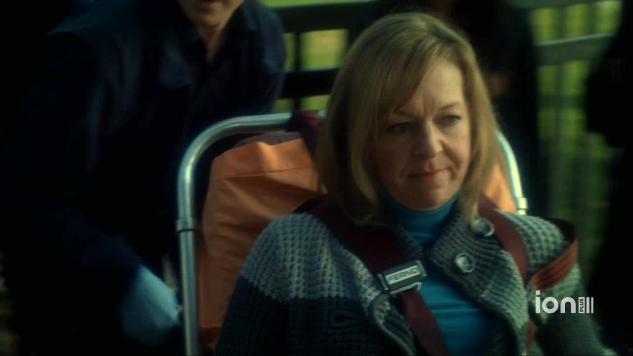 A woman in a blue turtleneck and patterned jacket sits strapped into a stretcher. Someone in a dark uniform is pushing the stretcher forward.
