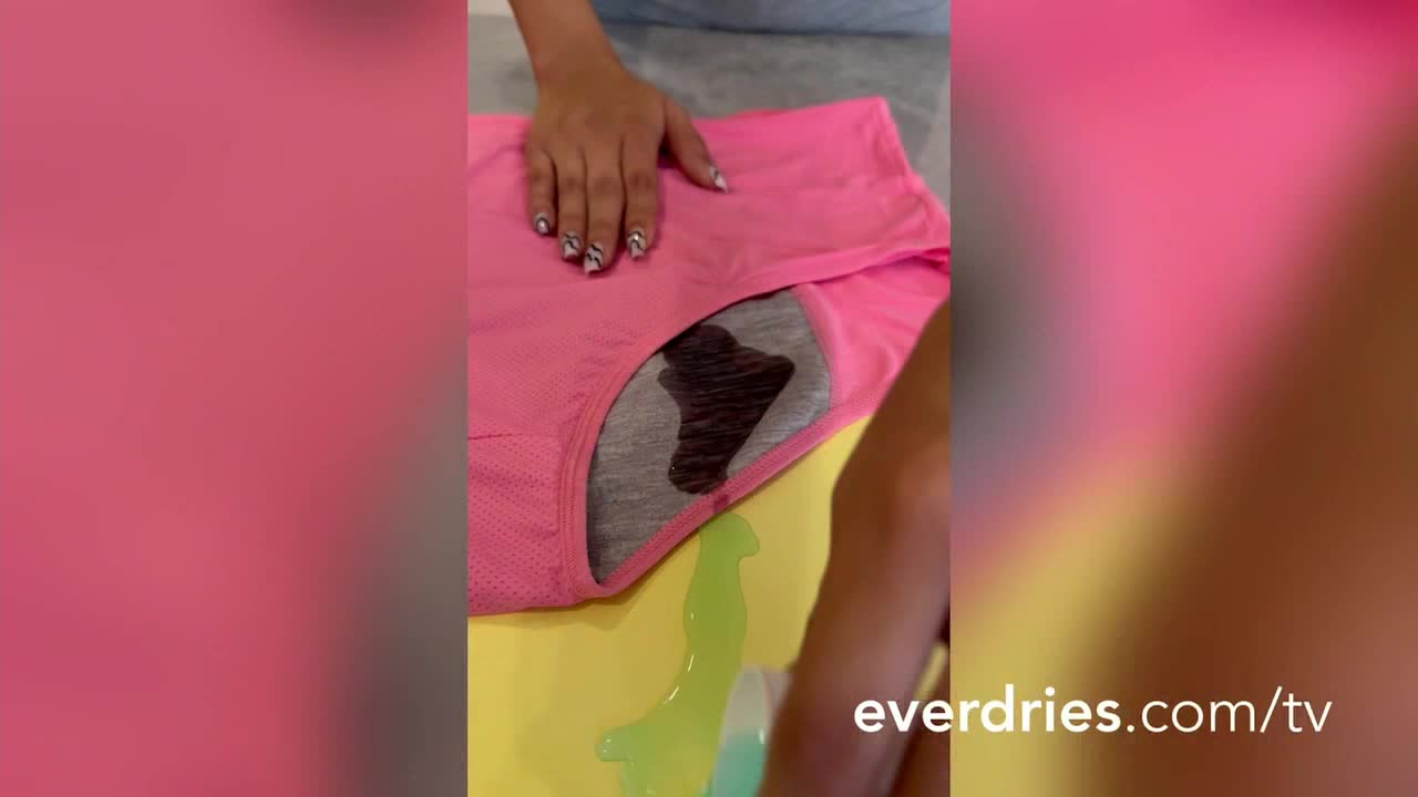 A pink garment sits on a yellow surface, stained with a dark liquid. A hand with manicured nails rests on the fabric, while a green liquid spreads across the surface.

