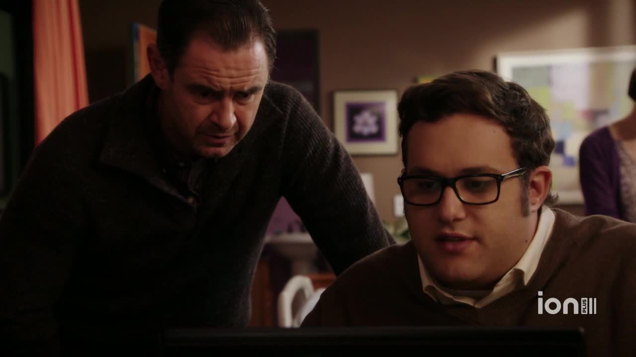 A man in a dark sweater leans over the shoulder of a younger man, both focused intently on a computer screen. The younger man, wearing glasses, appears to be speaking, while the older man listens with a furrowed brow.

