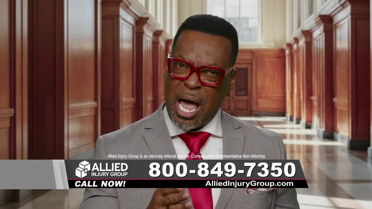 A man in a gray suit and bright red glasses is speaking directly to the camera, his mouth open. Below him, a graphic for Allied Injury Group flashes on the screen.
