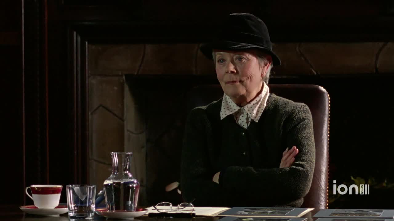 The woman in the hat sits behind a desk, arms crossed, looking directly ahead. A water pitcher and a teacup sit on the desk, next to some framed photographs.
