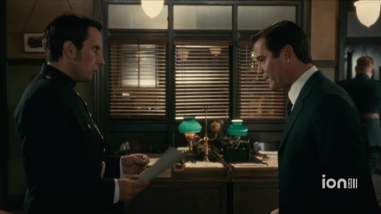 A uniformed officer holds a document, facing a man in a dark suit. The man in the suit leans in, listening intently in what appears to be an office.
