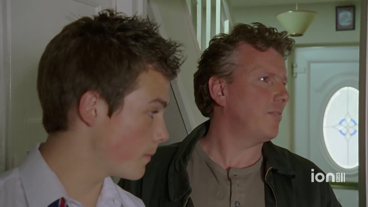 A man in a dark jacket is speaking, his gaze directed off-camera. Beside him, a younger man in a white shirt listens intently, his cheeks flushed.
