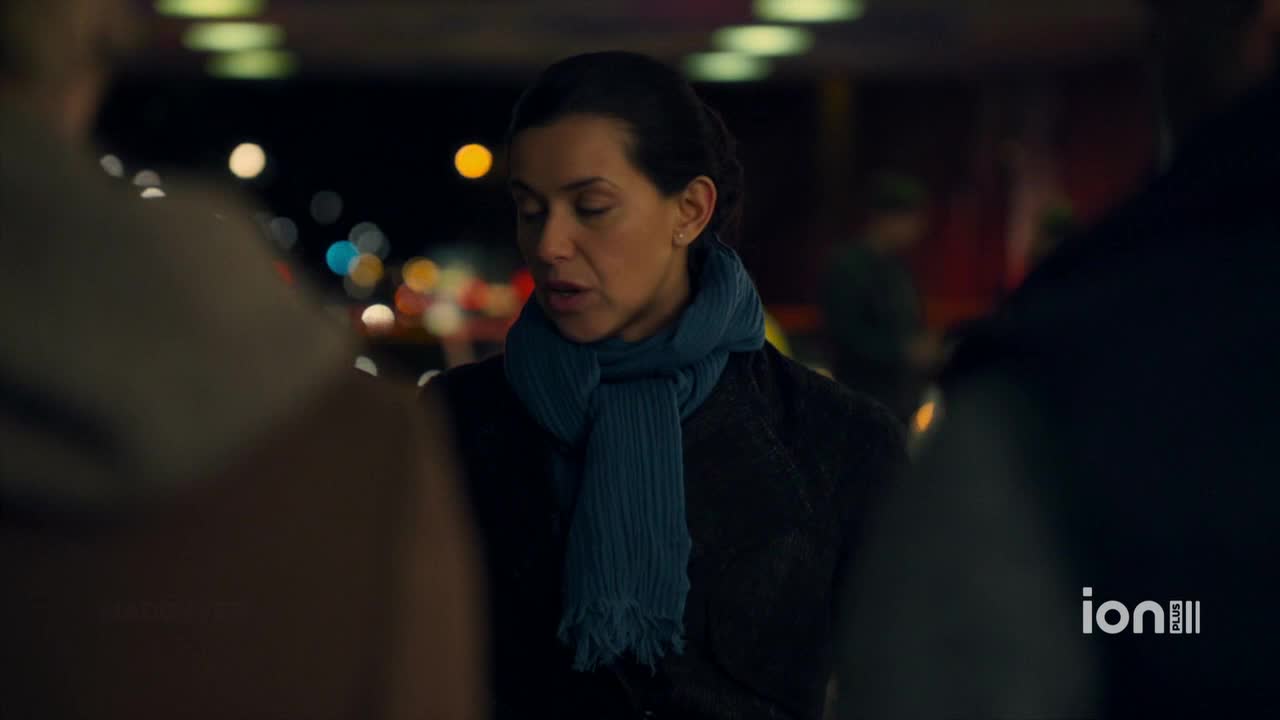 A woman in a blue scarf speaks, her gaze lowered as she addresses the people around her. The scene, lit by the lights of a city at night, is likely from a United States ION Plus show.
