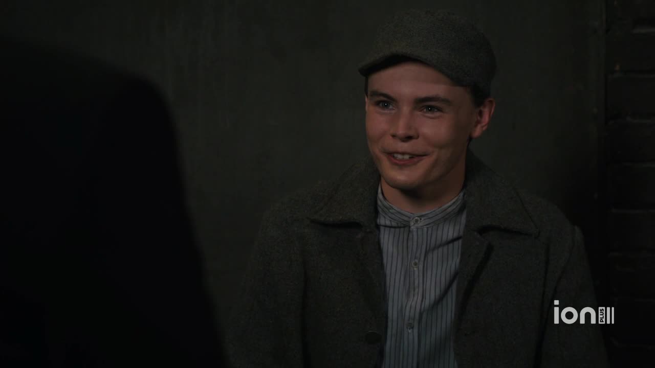 The young man in the cap smiles, his eyes crinkling at the corners. He's wearing a coat over a striped shirt, and seems to be listening intently.
