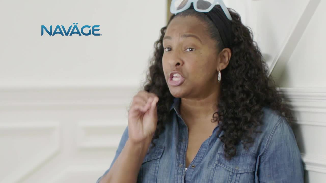 A woman in a denim shirt gestures with her right hand as she speaks, her mouth open. Her glasses are perched on her head, and she's standing in front of a wall.
