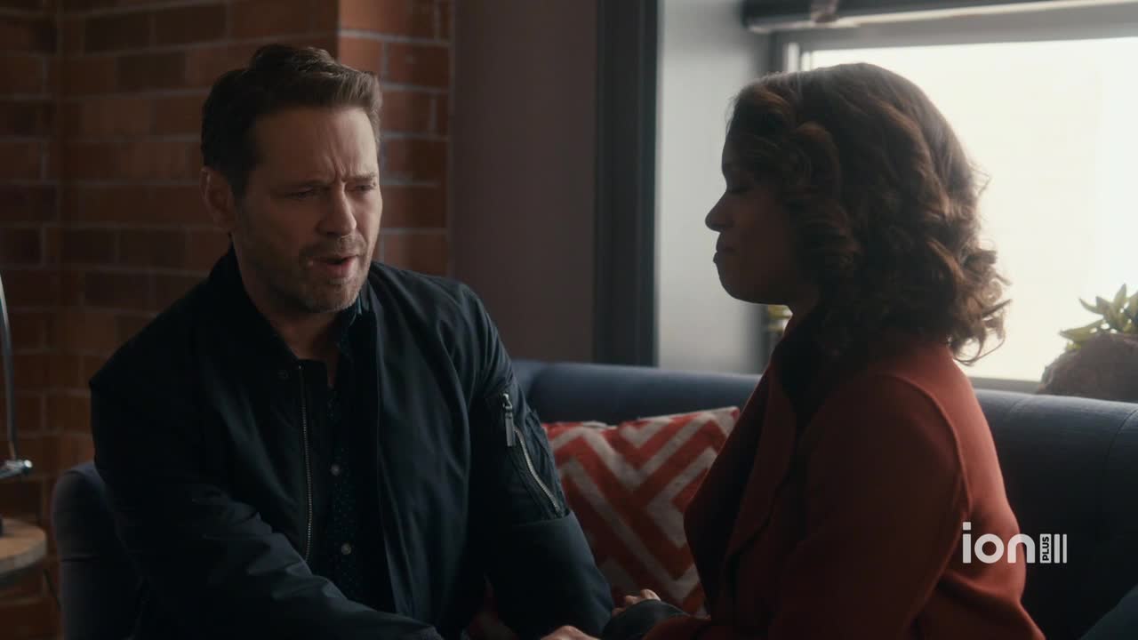 A man in a black jacket is speaking, his face etched with concern. Across from him, a woman in a red coat listens intently, her expression unreadable.

