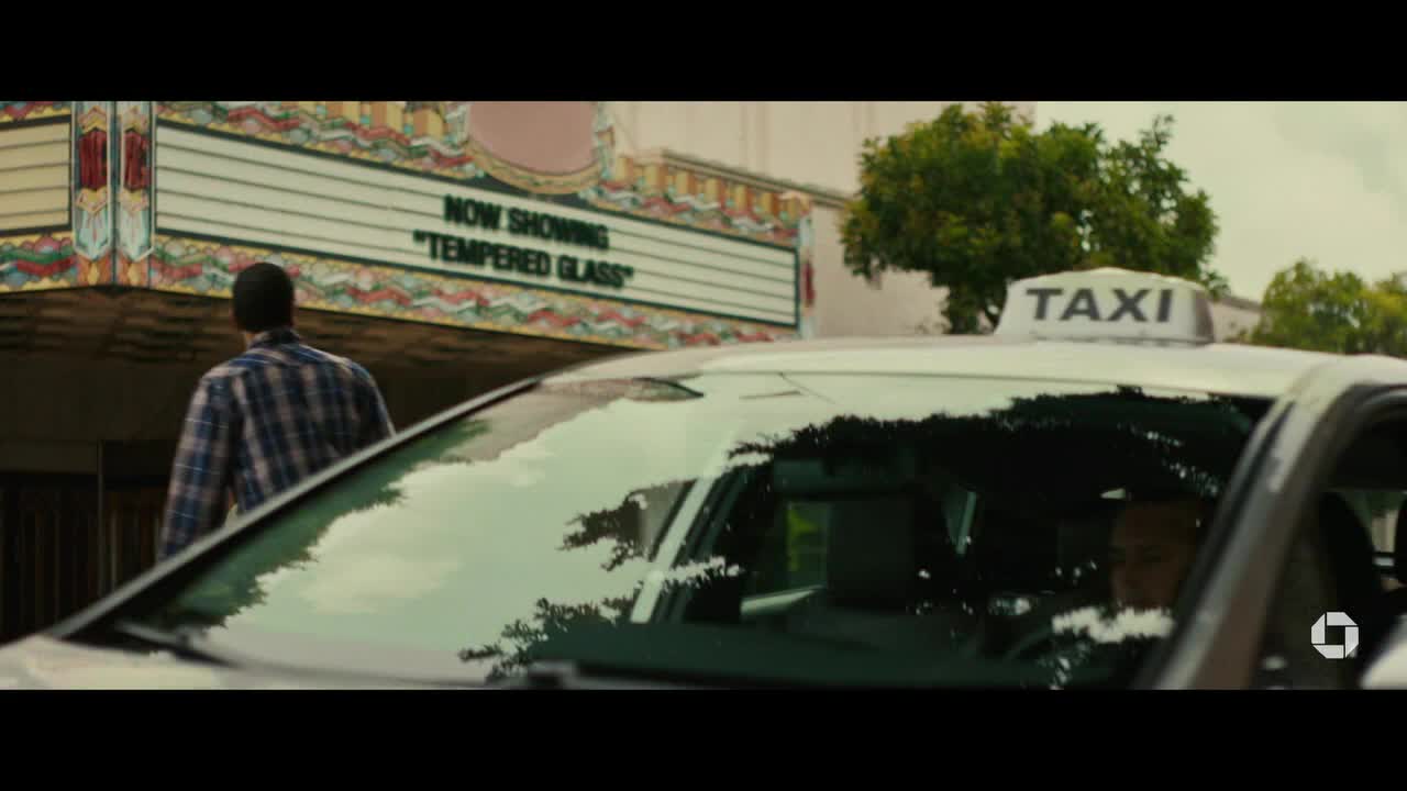 A man in a plaid shirt walks toward a movie theater, while a taxi sits idling nearby. The marquee advertises "Tempered Glass" as the current film.
