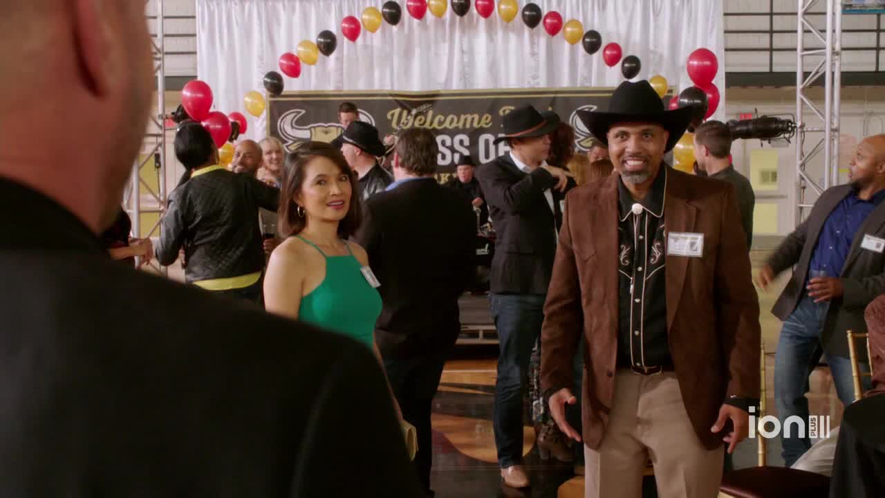 A man in a cowboy hat and brown jacket grins, gesturing with his hands. Guests mingle at a party with a "Welcome" banner and balloons overhead, likely from an ION Plus show.
