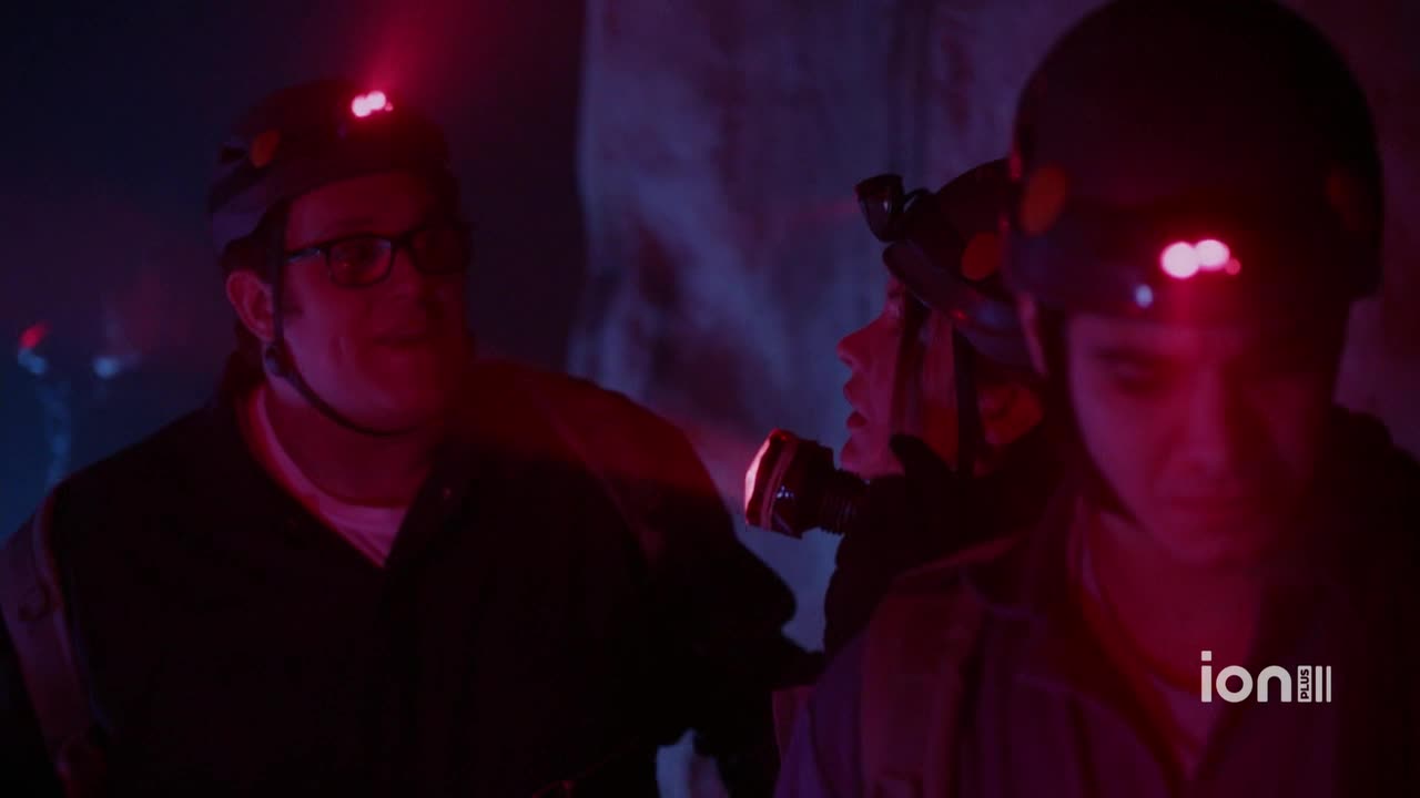Three people, all wearing helmets with red lights, stand close together in a dimly lit space. One man in the front wears glasses and seems to be looking off to the side.
