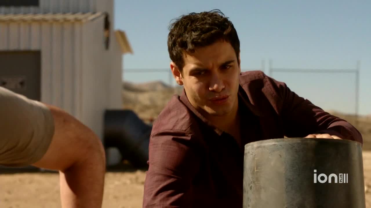 A man in a maroon shirt leans over a dark metal object, his gaze intense. The scene, likely from a show on ION Plus in the United States, is set in a dry, dusty area.
