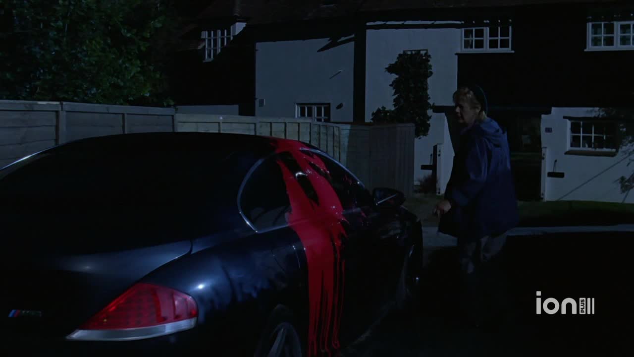 A dark car is splattered with red paint, streaks running down the side. A person in a blue coat walks away from the car, toward a house.
