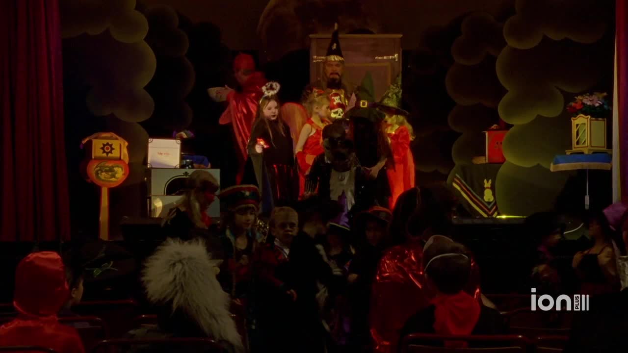 A group of kids in costumes are on stage, illuminated by stage lights. The ION Plus logo is in the corner, and the backdrop is decorated with oversized clouds.
