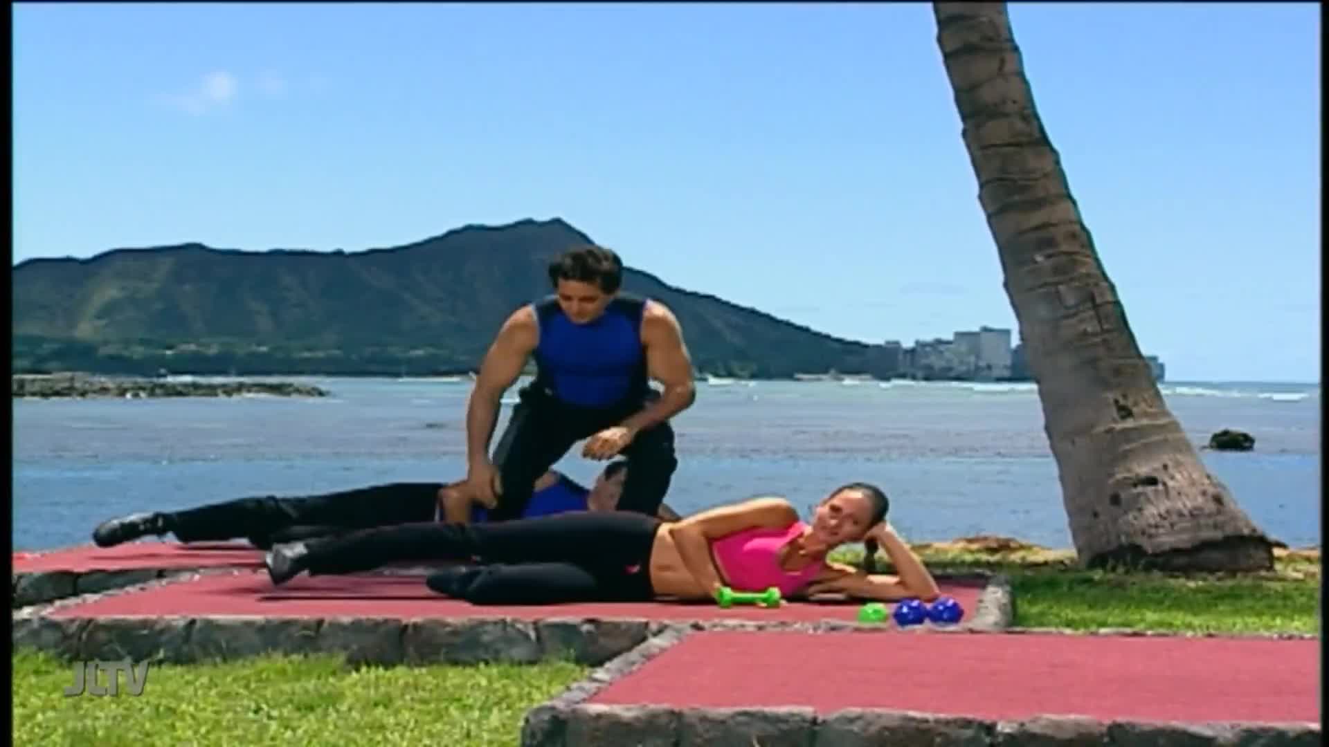 On a red mat by the water, two people are lying down while a man in a blue tank top assists one of them with an exercise. A woman in a pink top reclines nearby, dumbbells scattered in front of her.
