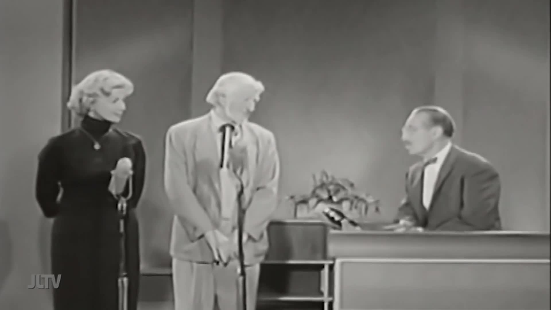 On the set of Jewish Life Television, a woman in a black dress stands next to a man in a light suit, both holding microphones. The host, in a dark suit and bow tie, gestures from behind a desk.
